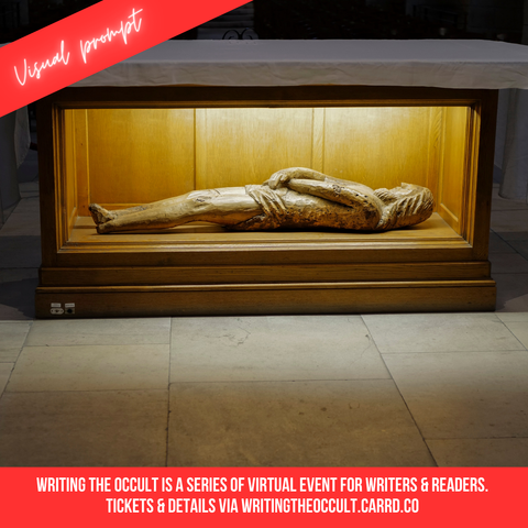 A wooden box with the camera-facing side in glass. Insight is a wooden carving of a human lying down with arms crossed across their belly. It is light with a spotlight, and the box has a white tablecloth draped over it.