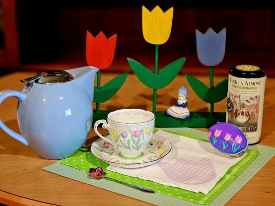 Tea tableau on a green ground with a white cloth appliqued with a lavender gingham tulip. Upon this are a vintage cup and saucer set patterned with a row of pastel tulips. Beside it are a handpainted purple rock with pink tulips, a tin of Vanilla Almond black tea, a powder blue teapot, and an iridescent purple spoon shaped like a flower. Behind them is a wooden sculpture of bright tulips in red, yellow, and blue, and a small Delft figurine of a little Dutch girl rests on the base.