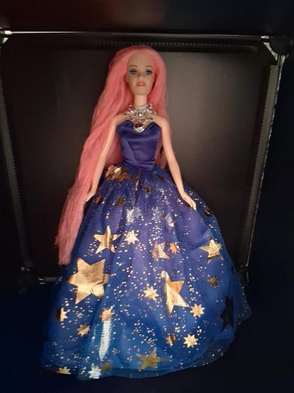 Una muñeca tipo Barbie. Es una gustomizacion hecha por mi. Es una Drag Queen y se llama Sasha Nova. Lleva el cabello rosa y largo y un maquillaje azulado con labios rosas acompañados de un elegante y mágico vestido azul eléctrico con un tul y estrellas doradas y brillantes por todo el vestido con un collar plateado