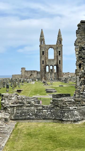 Portrait colour photograph of the ruined St Andrews Cathedral in Fife, Scotland. It was built in 1158 and became the centre of the Catholic Church in Scotland. Grass now covers the ground between the remaining walls and columns. The sea is just visible behind the site. The sky is blue, with high level cloud covering the majority of it.