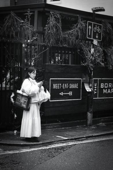 Black and white. A young woman with long black hair, in a long white dress and a short white padded winter jacket, with a woven Daunt Books shopping bag slung over her right shoulder and a white unfurled folding umbrella with the strap around her left wrist, is standing next to a Borough Market sign saying, "Meat, Eat, Share" while interacting with her smartphone.