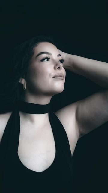 The image shows a woman with dark hair, wearing a black top. She has her arm raised, touching her head. The background is dark, highlighting her face and features.