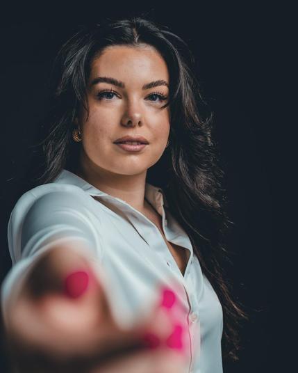 The image features a woman with long, dark hair, wearing a white shirt. Her focus is directed towards the camera, and her hand with pink painted nails is extended forward. The background is dark, highlighting her face and expression.