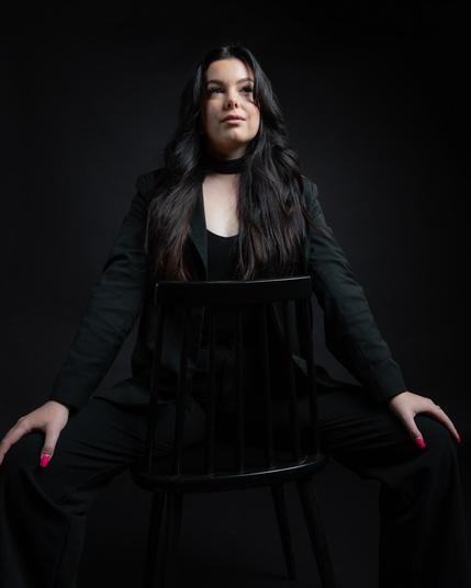 The image shows a person dressed in black sitting on a chair, posed in a confident manner. The background is dark, which puts the focus on the person in the image. Valokuvaaja Lappeenranta Kuvaajankulma