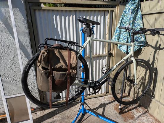 A gravel bike supported by a repair stand. There is a canvas pannier mounted on the stand.