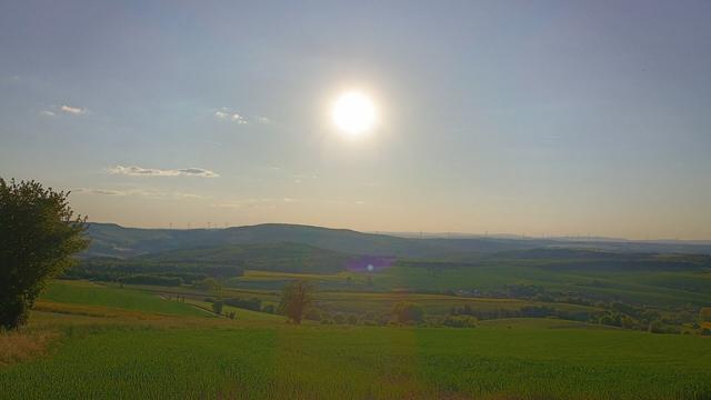 Blick über ein grünes Feld zu Hügeln am Horizont, darüber strahlt die Sonne.