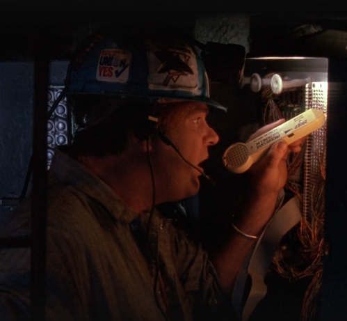 Screenshot of Dan Akroyd's character Mother, in 1992's "Sneakers," which is the single best hacking movie of all time.

In this picture, he's down inside a utility vault, wearing a call-center style boom mic headset under a blue hardhat.  He's using the receiver half of a  toner on a telephone system punch panel, while Whistler listens to the audio back in the van over the radio link.  They're identifying which copper pair is carrying the data signal they want to intercept by ear.

On the helmet is a beat up "UNION YES" sticker, next to a very large San Jose Sharks sticker.  The former is good social engineering practice; the latter is noteworthy because the Sharks were a new expansion team in the 91-92 season, so this was bleeding-edge Silicon Valley awareness.