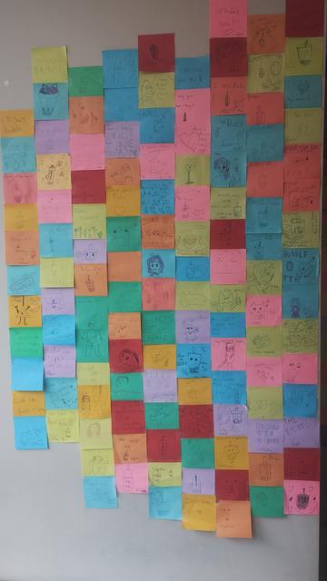 Multi-coloured mosaic of Post-It notes, each with doodles by children, covering the wall of the local bubble tea cafe