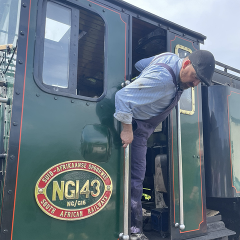 Train driver leaning out of engine cab with South African sign on engine