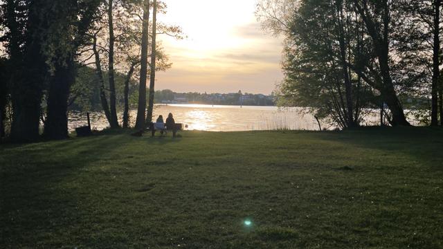 Setting sun in the upper center part, not clearly visible, sinking to clouds over a lake. The opposite side is visible with a small town panorama. Sun's blurred reflection on the calm water surface. The whole lower half of the photo is taken by a fine green lawn, with long shadows of the trees on each side. Small bench ideas visible by the water edge with two figures sitting on it and facing the sun.