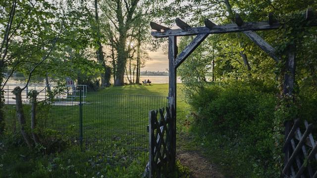 Open wooden gate with two wings from diagonally crossed pieces and a wooden rectangular arch above. Pathwat leads through it into the well kept park by the lake shore, fine lawn. Distant sun casting shadow.