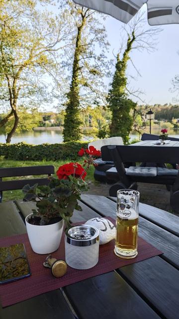 Restaurant garden table with half full beer glass, flower, ashtray and a room key. From the shadow area in the foreground, there's a view to the sunlit lake area with trees and houses on the opposite bank.