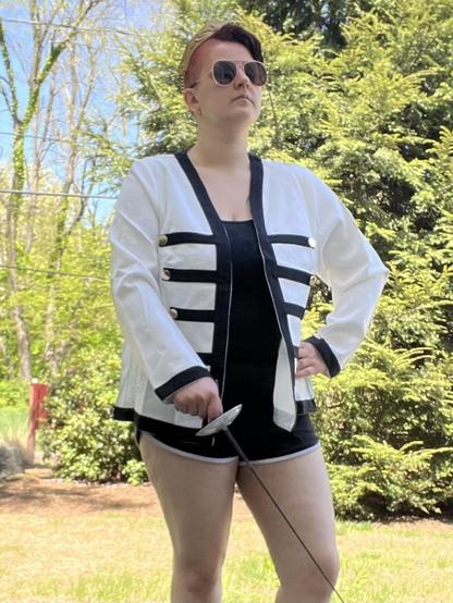 young white person (me). my short brown hair has been spray-dyed orange with cheap spray-on dye. i am wearing a white military/band style jacket, a plain black shirt underneath it, black booty shorts, sword earrings, a gold (and clearly handmade) crown, and aviator sunglasses. i am holding a fencing foil.