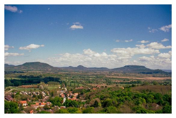 Color photograph of a Hungarian landscape with clouds. The photo was taken near Lake Balaton from a lookout tower with a view of the surrounding volcanoes. In the foreground you can see the houses of a small village. The blue sky, interspersed with a few clouds, extends to the center of the picture, where the volcanic hills can be seen on the horizon.