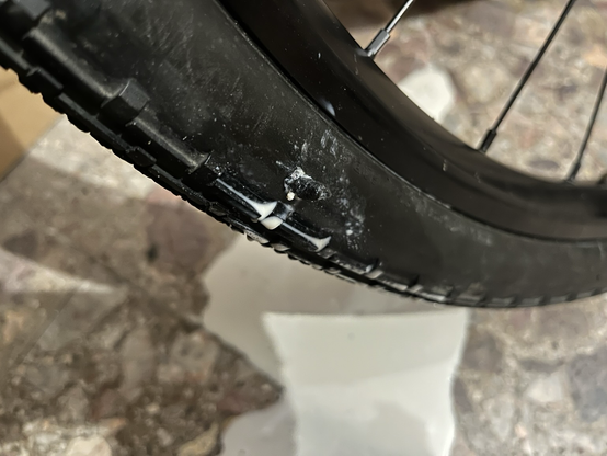 Closeup of the side of a tyre with a tubeless plug poking out and sealant spilled around the hole. A small pool of sealant on the floor in the background.