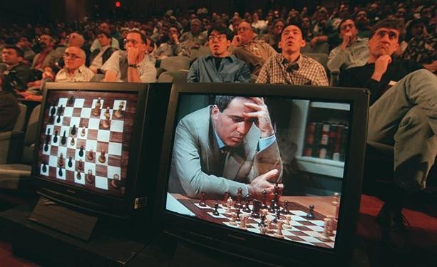 Zwei nebeneinander stehende Monitore in einem dunklen Saal: links eine digitale Schachbrett-Ansicht mit Figuren, rechts der Bildschirm, der den konzentriert nachdenkenden Schachspieler Kasparow im grauen Sakko mit der Hand an der Stirn zeigt. Im Hintergrund sitzt ein gespanntes Publikum in Reihen und verfolgt die Partie.