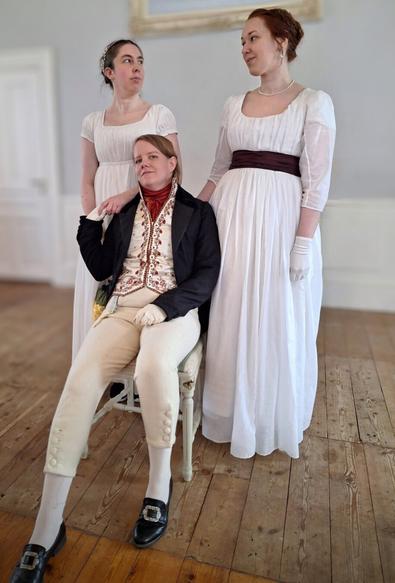 Alt text: Three people dressed in Regency-era clothing pose in a historical-style room with wooden floors and white walls. Two women stand wearing white empire-waist dresses, one with a dark sash, gloves, and pearl jewelry. Between them, a seated person wears cream breeches, a richly embroidered waistcoat, a dark jacket, and black shoes with buckles, striking a confident pose.