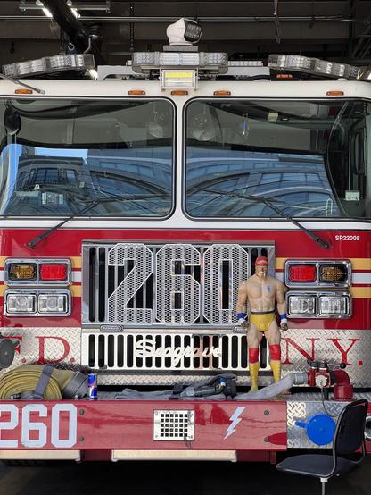 A fire truck is displayed with a prominent number "260" on the front. In front of the truck stands a figurine resembling a muscular man in wrestling attire, featuring yellow trunks and a red cap. The background shows industrial elements and additional fire truck