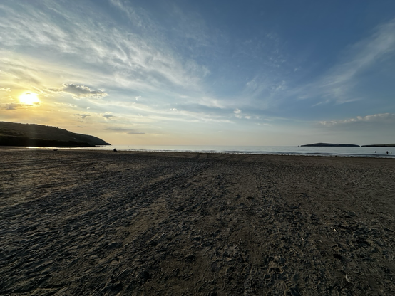 A tranquil beach at sunset, with the sun setting over the hills. The sky is painted with warm hues and soft clouds.