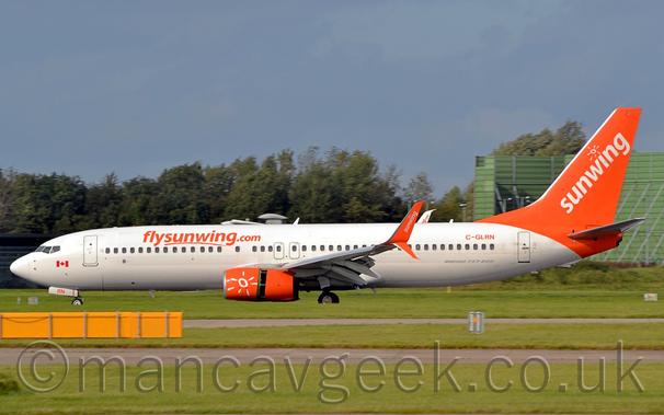 Side view of a white and orange, twin engined jet airliner moving from right to left.
The plane is mostly white, with orange "FlySunWing[dot]com" titles on the upper forward fuselage, and the orange registration "C-GLRN" on the upper rear.
The tail is a bright orange, with diagonal white "SunWing" titles under a white, hand-drawn sunburst logo.
This same logo is repeated on the orange engine pods under the wings.
The wingtips are split, turning both up and down, bright orange on the outer faces, and white on the inside, as can be seen on the other side of the plane.
The outer face has diagonal white "SunWing" titles, while the white inner face has orange titles.
The rear portion of the engine pods have moved rearward, exposing the thrust-reversers, allowing the plane to slow down rapidly.
Green grass separated by a runway and a taxiway fill the foreground, with trees and a large, green, box-like structure in the background, under grey-blue sky.