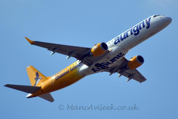Low side view of a white and yellow, twin engined jet airliner flying from left to right at a very low altitude with flaps extended from the rear of the wings, the undercarriage safely stowed away, and the nose raised sharply, suggesting it has just taken off.
The plane has a white forward fuselage, with blue billboard-style "Aurigny" titles, inverted and repeated under the belly.
The rear fuselage and tail are bright yellow, with an image of a flying puffin on the tail.
There are additional blue "Channel Islands" titles on the lower rear fuselage.
The engine pods under the wings are the same bright yellow, with a waving white flag with a red cross in the middle, overlaid with a smaller yellow cross.
Pale blue sky fills the rest of the frame.