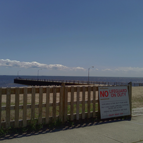 A photo from a beach scene. The clear sky shows a line of clouds running parallel with the horizon, as a short pier reaches towards them at an angle. An unpainted picket fence separates the sand from the foreground. A white sign indicates that there is no lifeguard on duty.