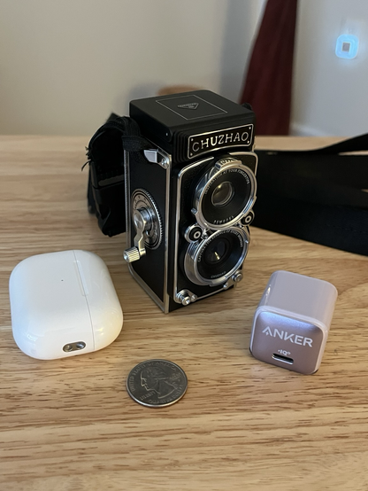 A closeup of the Chuzhao “TLR” toy camera, surrounded by an iPod case, a quarter, and a phone charger for size reference.