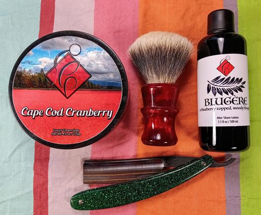 (Le Chat Mistral description)

The image showcases a collection of grooming items arranged on a striped background. On the left, there's a round tin of "Cape Cod Cranberry Luxury Cream Soap," featuring a scenic label with a red hue. In the centre, a red shaving brush with white bristles stands upright. To the right, there's a dark bottle of "Blugère," a blueberry-topped, woody fougère aftershave lotion. Below these items, a green and silver straight razor with a glittery handle lies horizontally. The items are neatly organised, suggesting a traditional shaving setup.