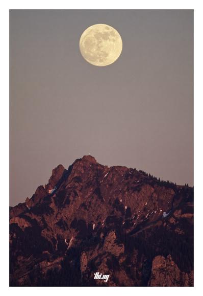 Photo of an almost full moon above a rocky mountain peak and surrounding cliffs, all illuminated by intense alpenglow, the last light of the day/tonight. Clear sky.