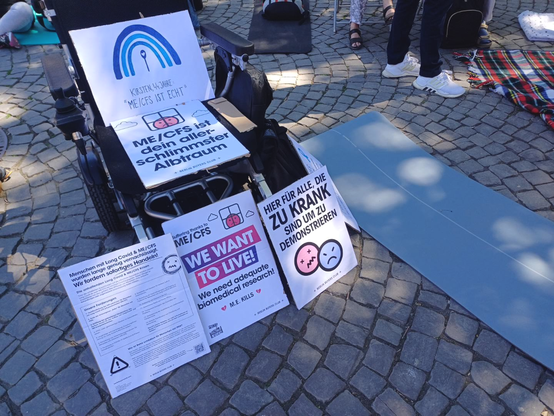 an einem Rollstuhl stehen und liegen mehrere Schilder zum Thema chronische Müdigkeit, wie "ME/CFS ist dein schlimmser Alptraum", "We want to live" und "Hier für alle, die zu krank sind um zu demonstrieren", daneben liegen Decken und Matten auf dem Straßenpflaster