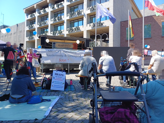 Demonstrant*innen sitzen auf Decken und Bänken vor einer mit weißen und blauen Luftballons geschmückten Bühne mit einem Banner "ME/CFS - Forschung, Anerkennung und Versorgung jetzt!"; im Hintergrund das Rathaus in der Kölner Altstadt (mit Flaggen von Köln, Deutschland und EU); im Vordergrund steht ein Rollator
