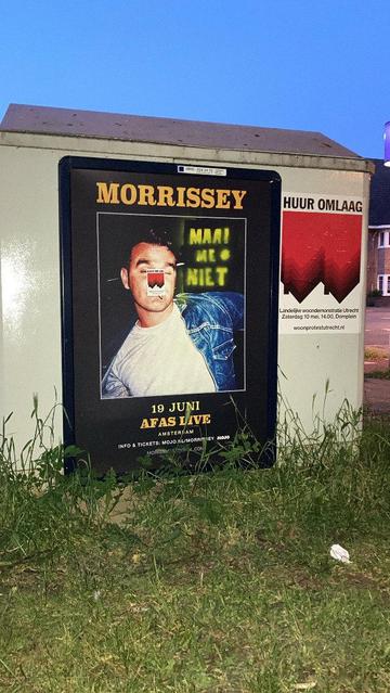 reclamebord op kastje. op het bord is in geel maai me niet * gesprayed. Naast het bord hangt een huur omlaag poster.
De reclame is een aankondiging van concert. Op de neus van de artiest zit een woon omlaag sticker. Gras voor, blauwe lucht