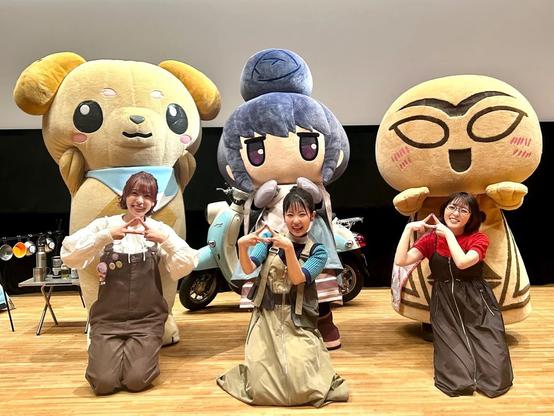 Three women kneel on a stage in front of large, colorful character mascots, all smiling and making a hand gesture resembling a triangle. Behind them are oversized plush characters, including a dog, a blue-haired girl, and a tan creature with large glasses and pink cheeks.