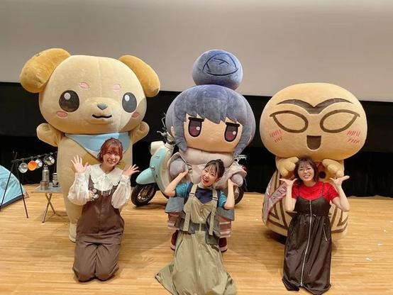 Three women kneel on a stage in front of large, colorful character mascots, all smiling and making a hand gesture resembling a triangle. Behind them are oversized plush characters, including a dog, a blue-haired girl, and a tan creature with large glasses and pink cheeks.