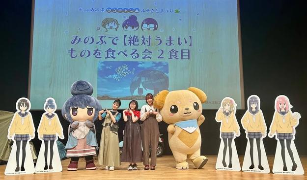 This image shows a stage event featuring three women posing with life-sized character mascots and cardboard standees from the anime Yuru Camp (Laid-Back Camp). Behind them is a large screen with event details in Japanese, celebrating food and the show’s setting in Minobu. The cheerful atmosphere and themed characters suggest a fan gathering or promotional celebration tied to the series.