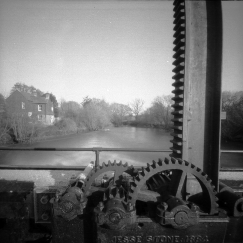 Black and white photograph looking down a river from the bridge above a weir. In the foreground are the cast iron gears for the machinery to open the sluice gates.