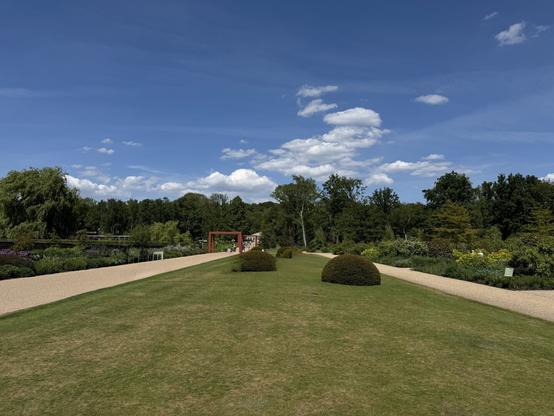 An open expanse of lawn, with a gravel path on both sides. Trees and bushes in the distance.