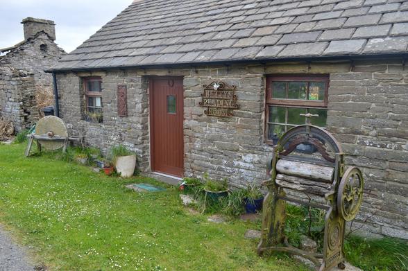 The exterior of a small single-storey stone cottage with a metal sign by the door 'Betty's Reading Room' . An old metal mangle and various random bits and bobs outside, a small patch of grass speckled with daisies, a low slate roof.