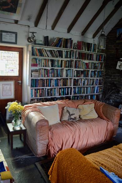 The interior of the space, lined with books. There's a comfy old sofa, a low beamed roof, and some fresh flowers in a vase. Out of sight, there's a small fire and yet more books. An old stone floor, little coffee tables laden with things to read, squished cushions and a sense of a much-loved space.