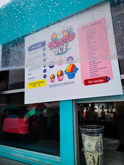 A color photo of a shaved ice stand. The menu  at top is partially obscured by raindrops on a clear umbrella. There is a plastic tip jar in the front with a dollar bill visible. People are dimly reflected in the glass and in the back are bottles of flavor syrup.
