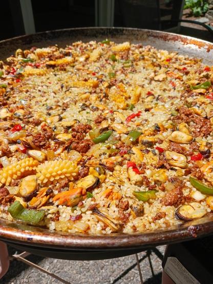 Photo of a large shallow pan - sides cut out of the frame - of paella cooking outdoors. The pan is a mix of rice, ground chorizo, chopped onions, chopped green and red bell peppers, "flower cut" calamari, and mussels.