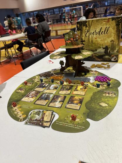 Foto do jogo Everdell montado em cima de uma mesa no SESC. O tabuleiro é verde com uma árvore de papelão em cima que foram duas plataformas onde se posicionam peças e cartas. Em cima do tabuleiro, pecinhas de recursos, cartas e peças do jogador
