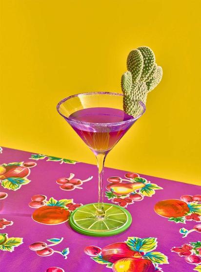 A purple cocktail in a martini glass with a cactus-shaped garnish, set on a colorful fruity tablecloth against a bright yellow background.