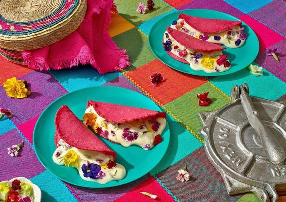 Three bright pink tacos filled with creamy white filling and colorful edible flowers on turquoise plates, set on a vibrant checkered tablecloth with a tortilla press and woven hats nearby.