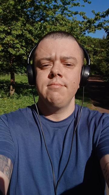 A white transmasc wearing a blue t shirt and black headphones. He has a buzzed head and is stood in a park