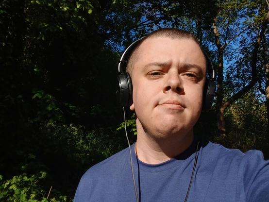 A white transmasc wearing a blue t shirt and black headphones. He has a buzzed head and is stood in a park