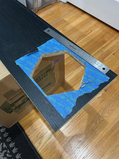 A black-wrapped particle IKEA BILLY shelf with a large hexagonal hole cut through it.  The area around the hole is covered in painter's tape.  A metal ruler is nearby for scale.