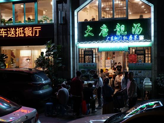 Neon signs light up the front of two restaurants in Shenzhen, China. People mill around outside of the restaurant on the right waiting to get in
