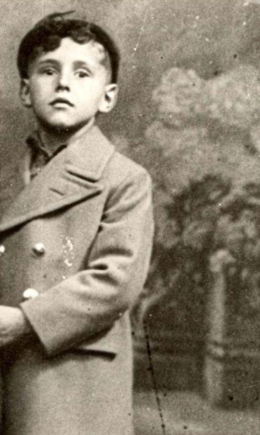 A young boy in a cap and coat photograph against a studio background with some fence and flowers.