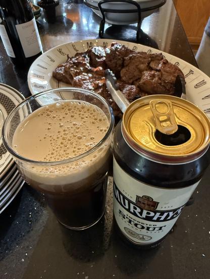 A pint can of Murphys stout beer and a pint glass with a thick tan head are in front of platter full of cooked steaks